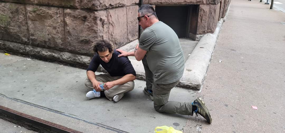A Semper Fi Mission volunteer kneeling to help a man seated on a city sidewalk